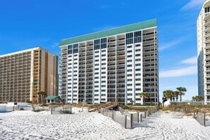 A beachfront hotel with multiple floors, a green roof, and a sandy beach area with wooden fences. - Emerald Towers #205 OnTheGulf*LotsOfResortAmenites (DESTIN)