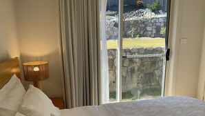 Ein Schlafzimmer mit einem Bett, einer Lampe, Vorhängen und Blick auf eine Steinsäule sowie Vegetation draußen.