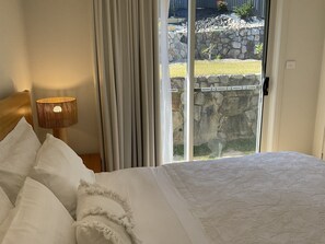 Slaapkamer met een bed, een lamp, gordijnen en uitzicht op een stenen muur en begroeiing.