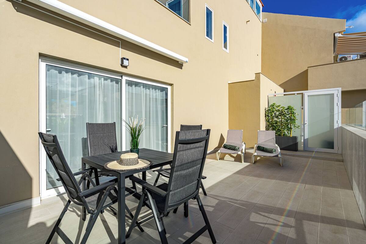 Un patio exterior con una mesa y sillas, un ventanal corredero y un pequeño rincón de descanso con una planta.