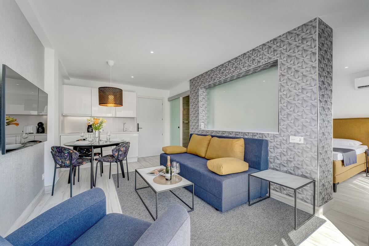 Una sala de estar moderna con sofá, mesa de centro y sillas. Hay un comedor con mesa y sillas. La habitación tiene un ventanal y una pared con diseño.