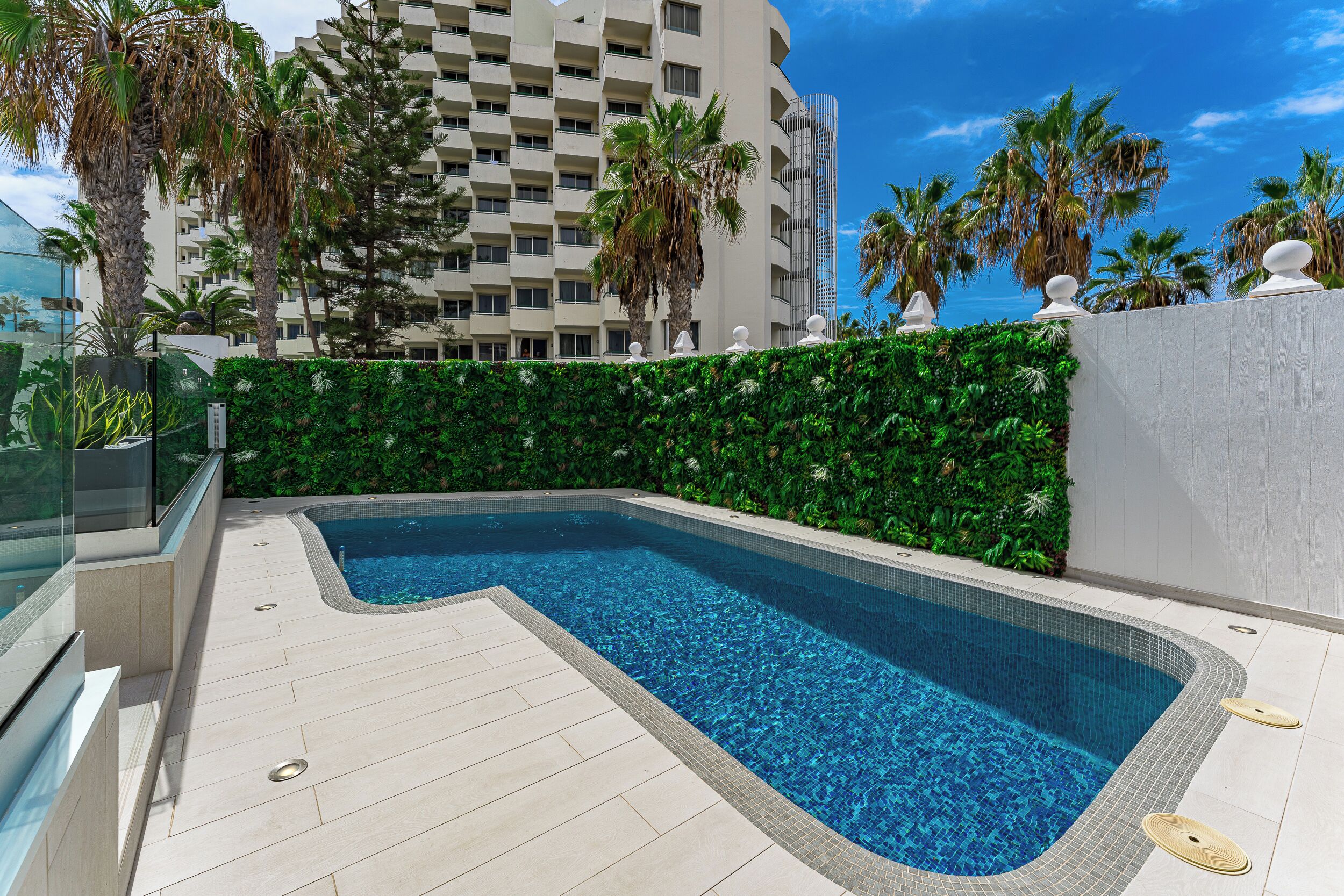 Una piscina con solárium empinado, rodeada de follaje y palmeras, con un edificio alto al fondo.