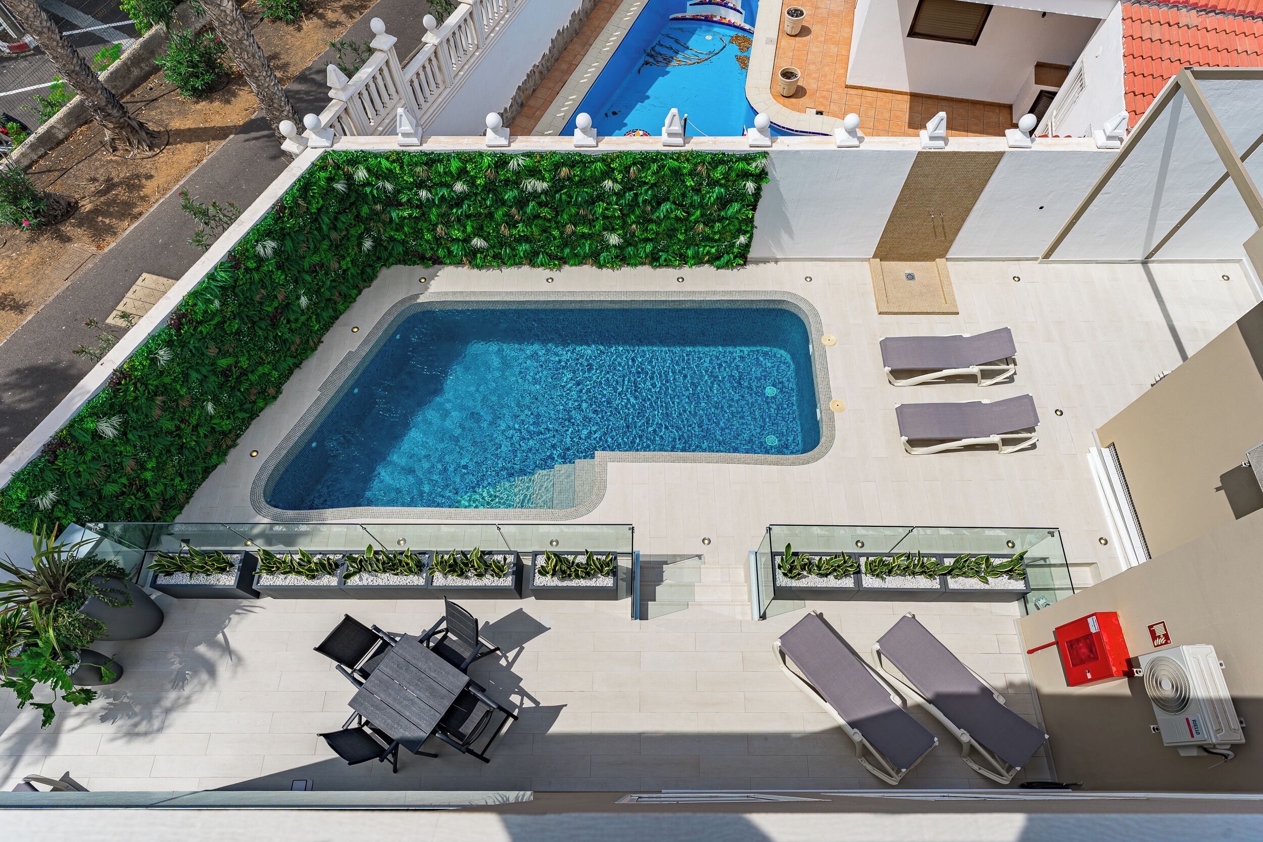 An aerial view of a rooftop swimming pool with lounge chairs and a table, surrounded by greenery and a white fence.