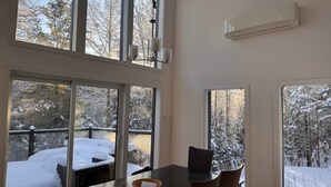 Un coin repas avec une table et des chaises, de grandes fenêtres donnant sur une terrasse enneigée et des arbres, et un appareil de climatisation fixé au mur.