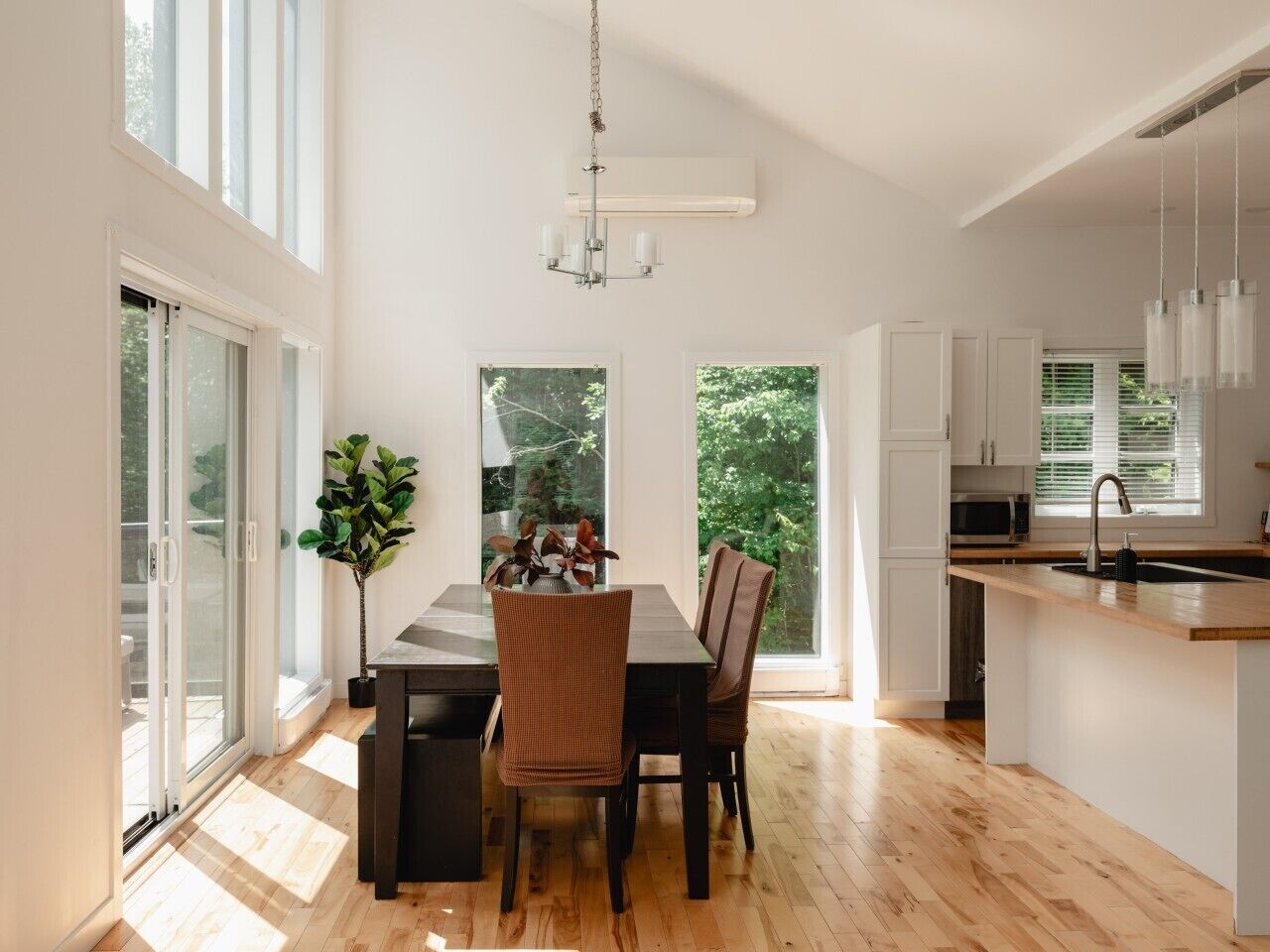 A modern kitchen with a dining area, large windows, and a view of greenery.
