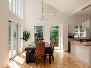 A modern kitchen with a dining area, large windows, and a view of greenery.