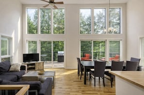 Un salon spacieux équipé d’un téléviseur, d’un canapé, d’une table à manger, de chaises et de grandes fenêtres donnant sur une terrasse avec un barbecue.