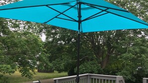 A deck with a glass-top table and chairs, surrounded by a wooden deck and a blue umbrella. - Ocean view 5-bedroom house in Narragansett with AC, WiFi, beach pass (Narragansett)