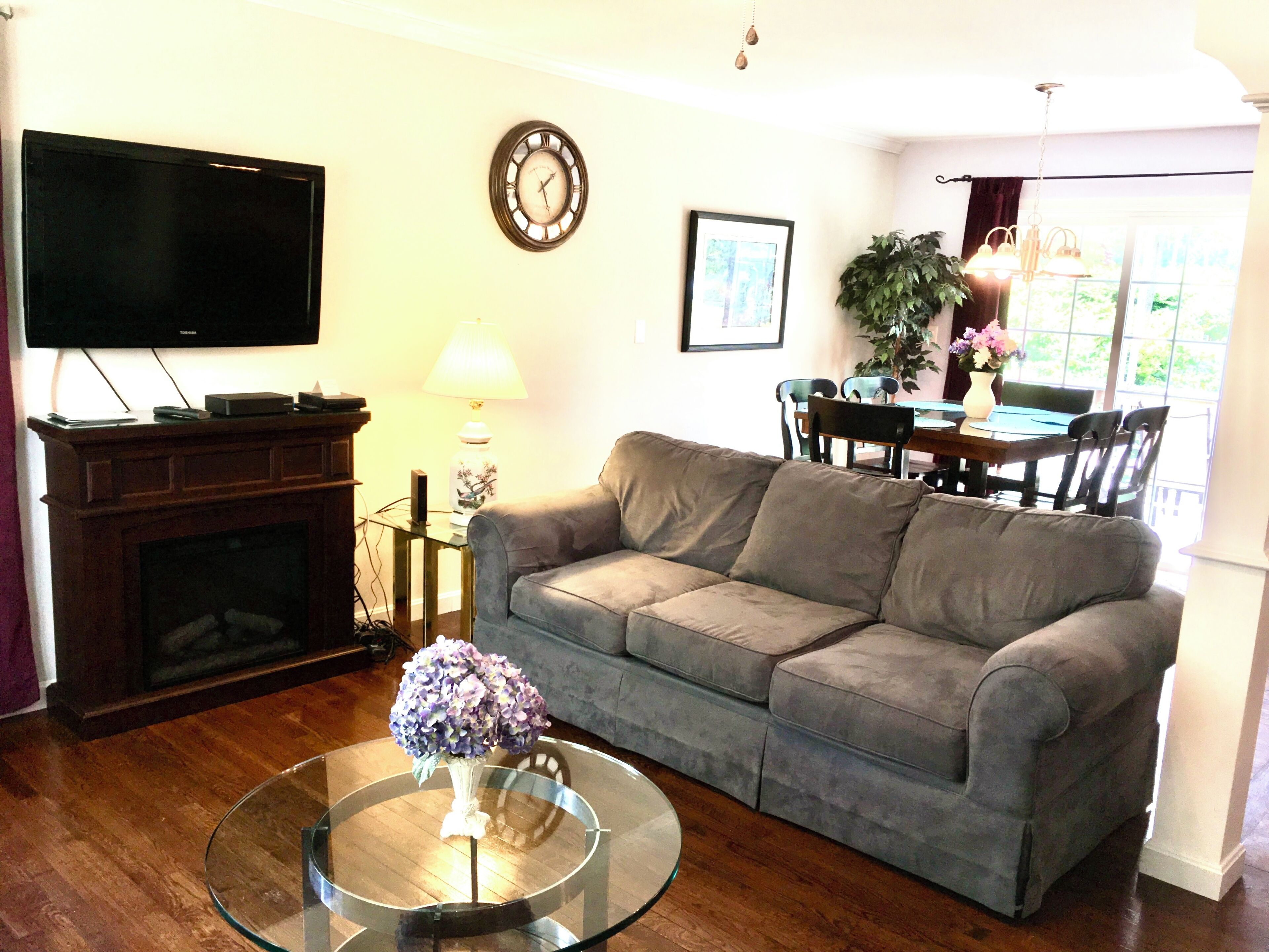 Una sala de estar con un sofá gris, una mesa de centro de vidrio con un florero, una chimenea, un televisor, una mesa de comedor con sillas y un reloj en la pared.