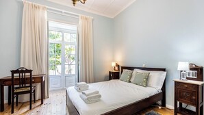 A bedroom with a wooden bed, a desk, and a chair. There is a window with curtains, a chandelier, and a nightstand with a lamp. - Casa Matias - Stylish 4BR Apartment • Lagos Old Town (Lagos)