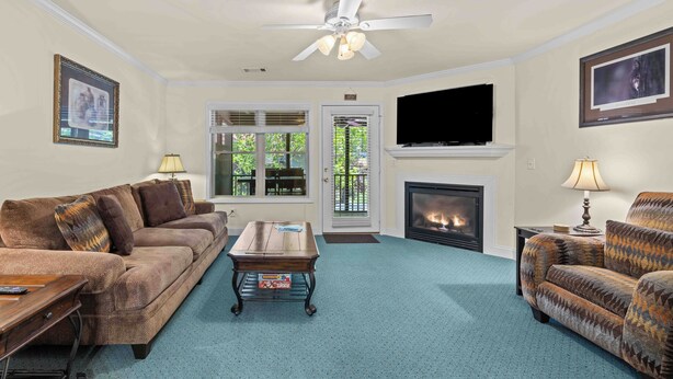 A living room with a sofa, a coffee table, a fireplace, a television, and a ceiling fan. - Uptown Getaway - Lovely Condo in Downtown Tuscany Villas (Helen)