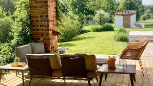 Une terrasse extérieure avec une plateforme en bois, un pilier en briques, des meubles en osier et une vue sur un jardin et un panneau solaire.