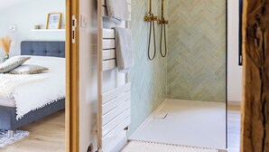 Une salle de bain moderne avec une douche en verre, un plancher en bois et un lit visible dans la pièce adjacente, avec une tête de lit grise.