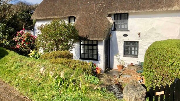 Une maison de campagne traditionnelle à toit de chaume, avec une façade blanche, un petit porche à l’avant et un jardin bien entretenu.