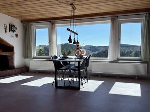 Un'area pranzo con tavolo e sedie, ampie finestre che offrono vista su un paesaggio boschivo e un camino con rivestimento in mattoni.