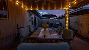 An outdoor patio with a table and chairs, a vase with sunflowers, string lights, and a grill in the background.