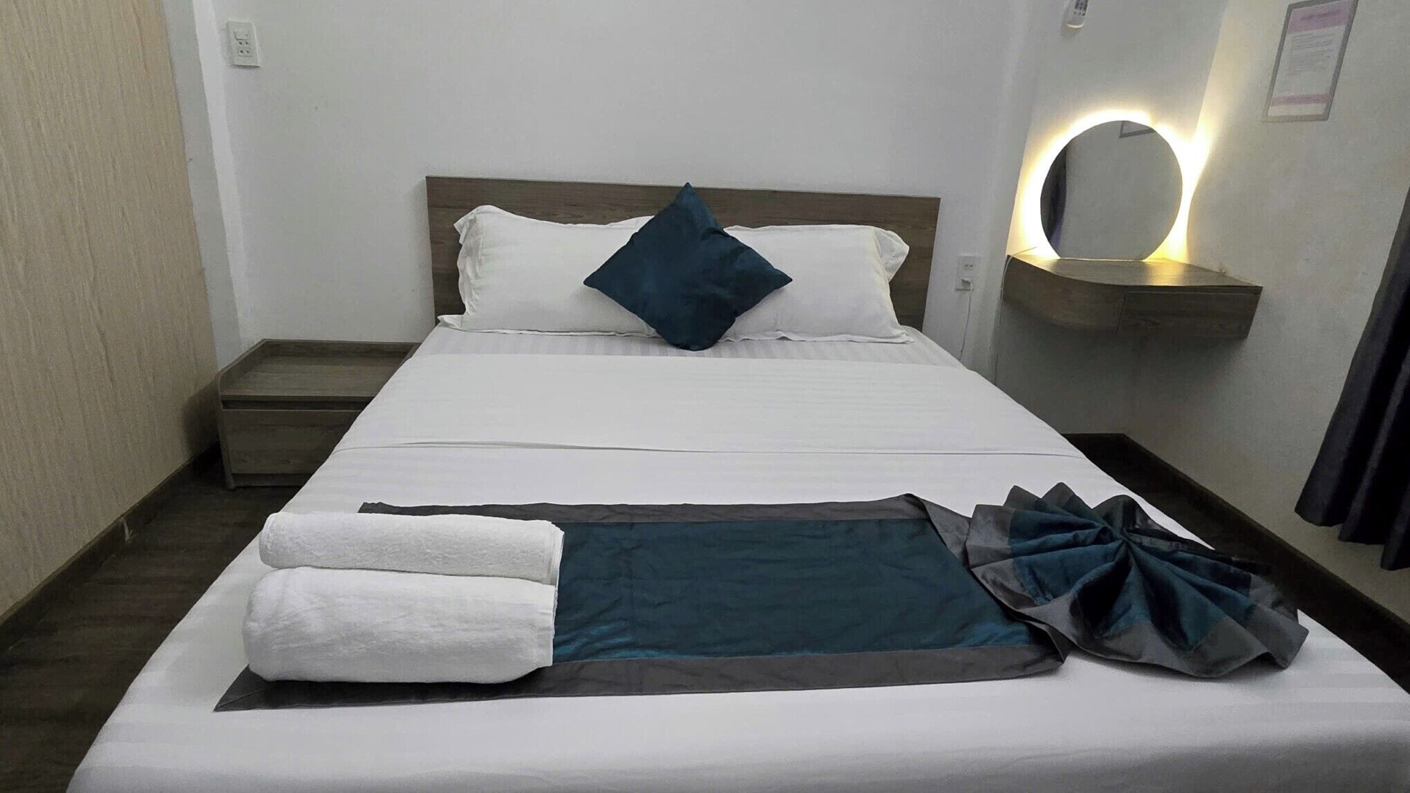 A neatly made bed with pillows, a folded blanket, and a towel. There is a bedside table with a lamp and a mirror.