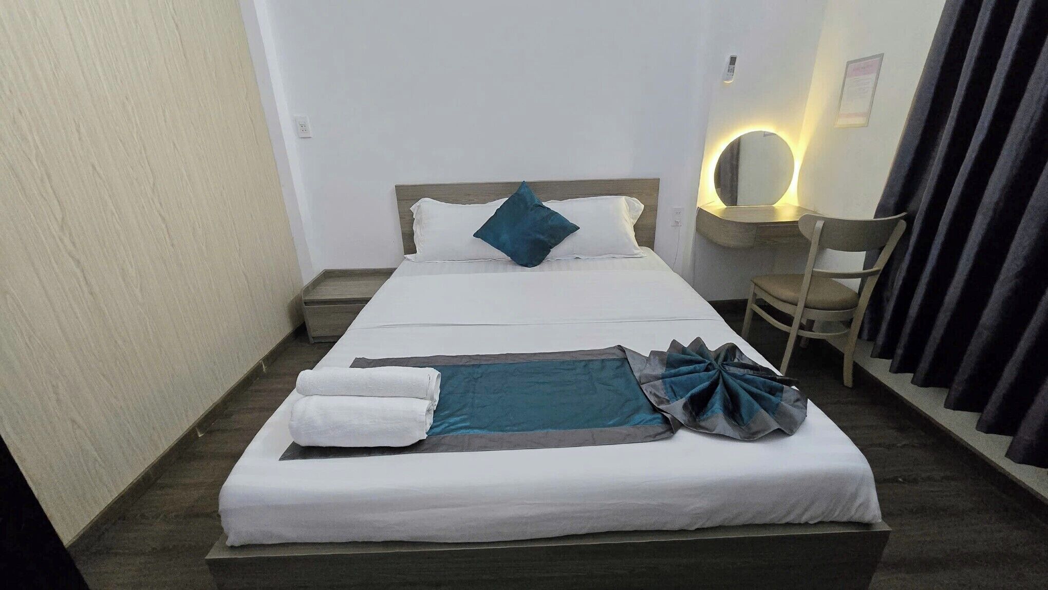 Una cama bien hecha con almohadas y una manta, una mesita de noche con una lámpara y una silla con cortinas.