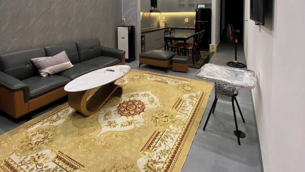 Un soggiorno moderno con divano, tavolino e tappeto, una cucina sullo sfondo e una TV a parete.
