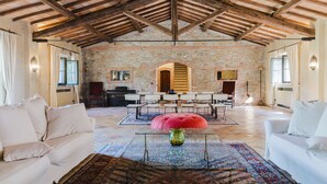 A spacious room with a high wooden ceiling, stone walls, and a large window. There are two white sofas, a red ottoman, and a glass coffee table with a vase. - Villa San Girolamo (Toscana)