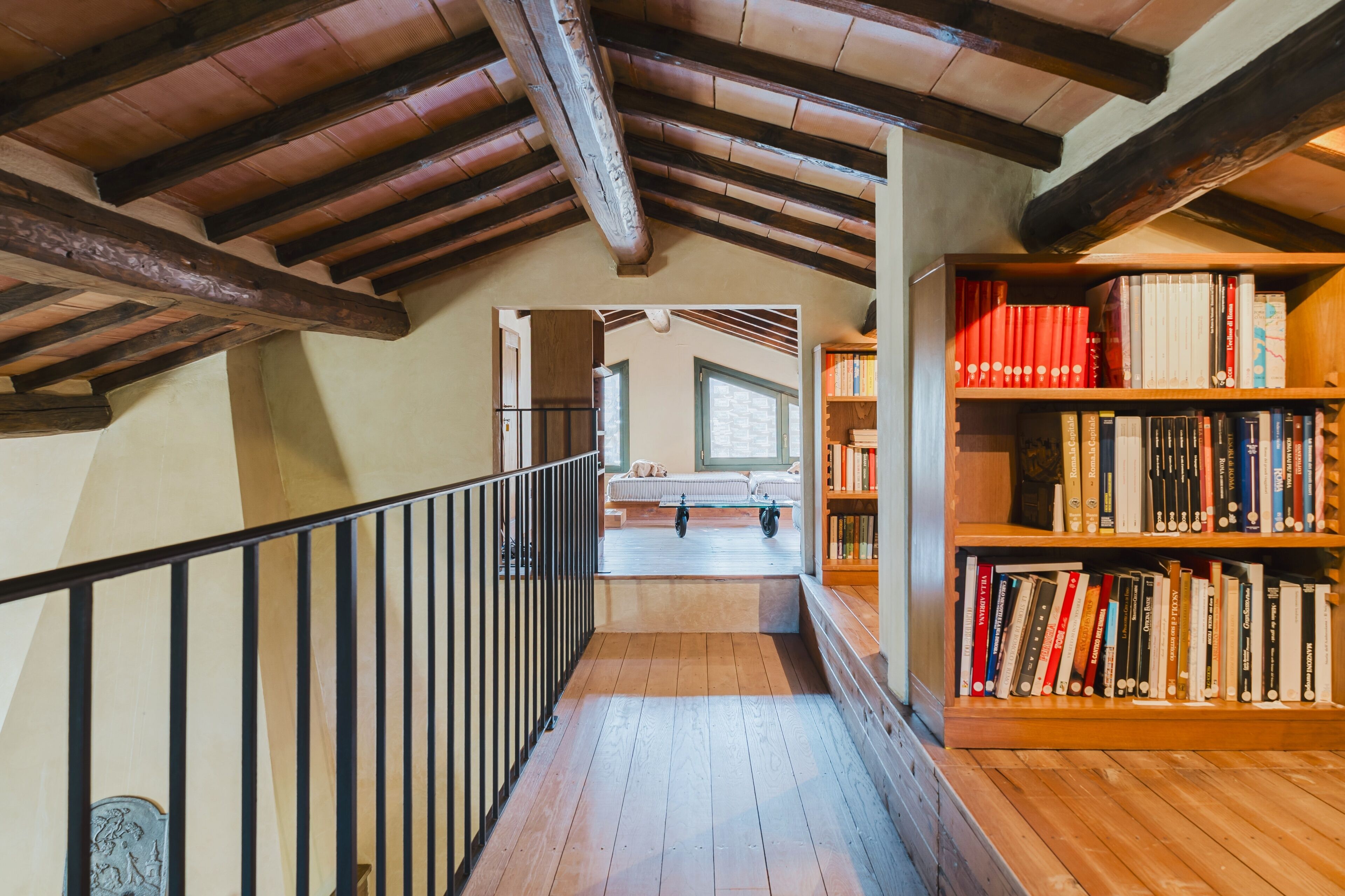 Eine Holztreppe führt zu einem Schlafzimmer auf dem Dachboden, das ein geneigtes Dach und freiliegende Deckenbalken hat. Rechts ist ein Bücherregal mit Büchern zu sehen.