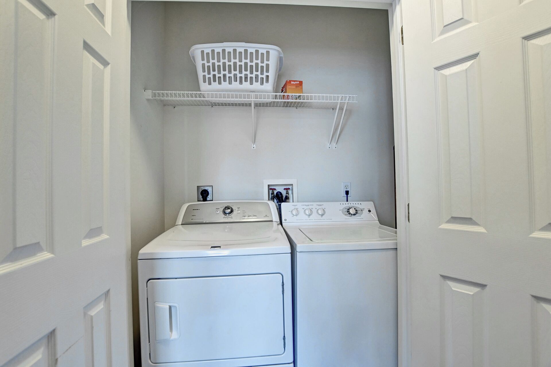 A laundry room with two stacked washers and dryers, a white laundry basket on a shelf, and a power outlet on the wall.