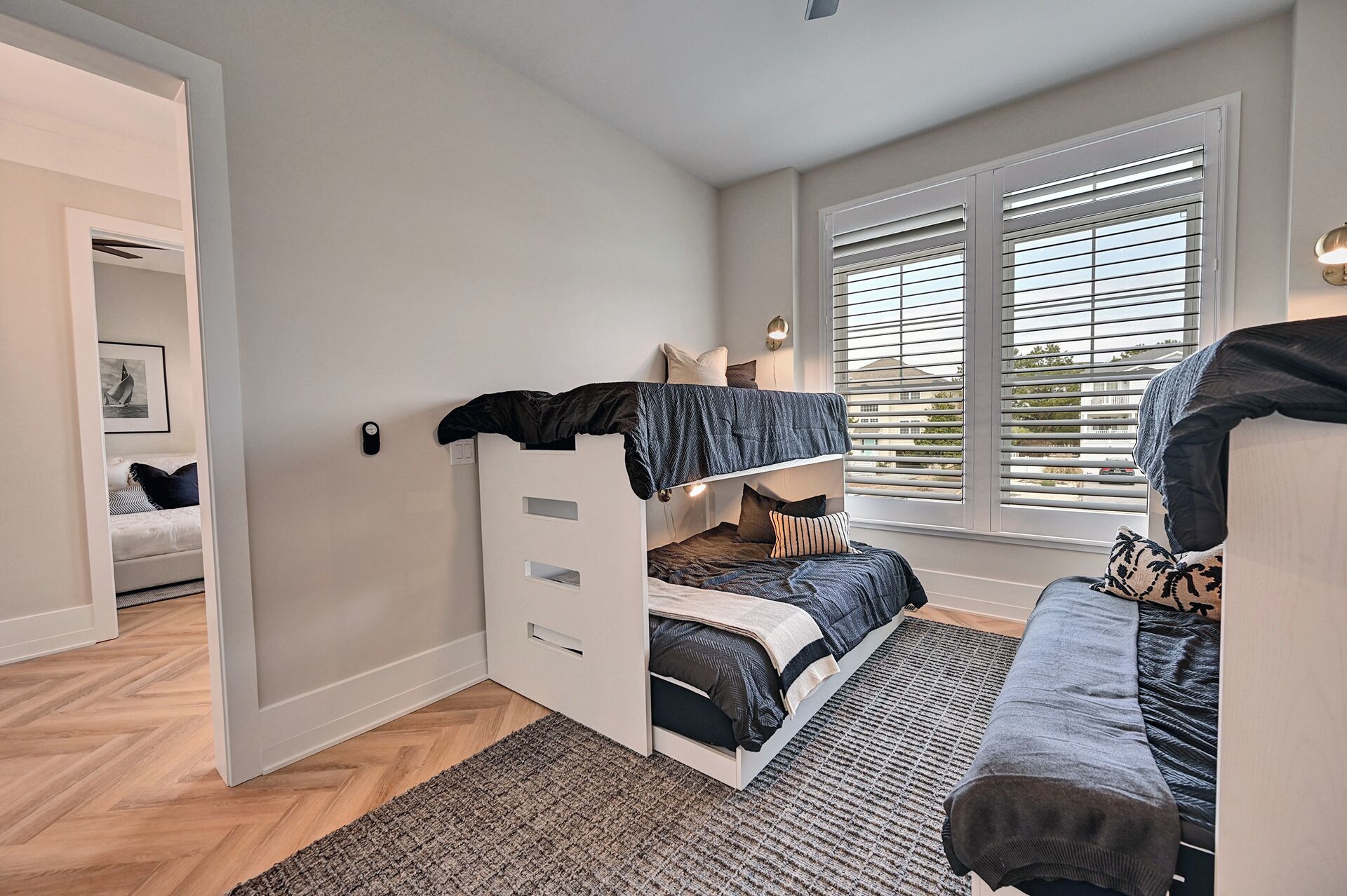 A room with a bunk bed, a sofa, a herringbone-patterned rug, and a window with blinds.