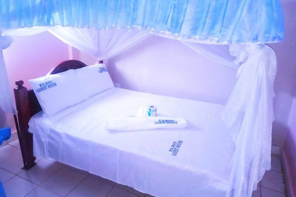 Een eenpersoonsbed met hemelbed, witte beddengoed en kussens met de tekst 'ELRIS GUEST HOUSE'.