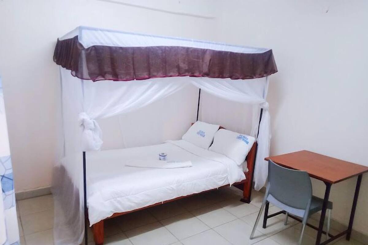 Een eenpersoonsbed met hemelbed, een tafeltje en een stoel in een kamer met witte wanden en betegelde vloer.