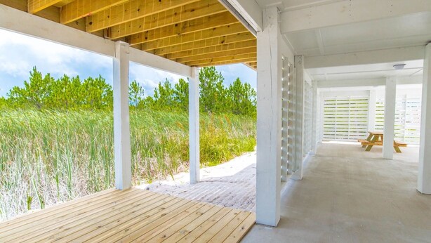 An open-air covered area with a wooden deck, white columns, and a view of a grassy landscape.