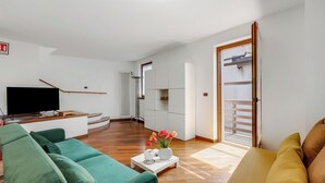A modern living room with a green sofa, a yellow sofa, and a coffee table with a vase of flowers. There is a flat-screen TV on a wooden stand, a radiator, and a sliding glass door leading to a balcony.