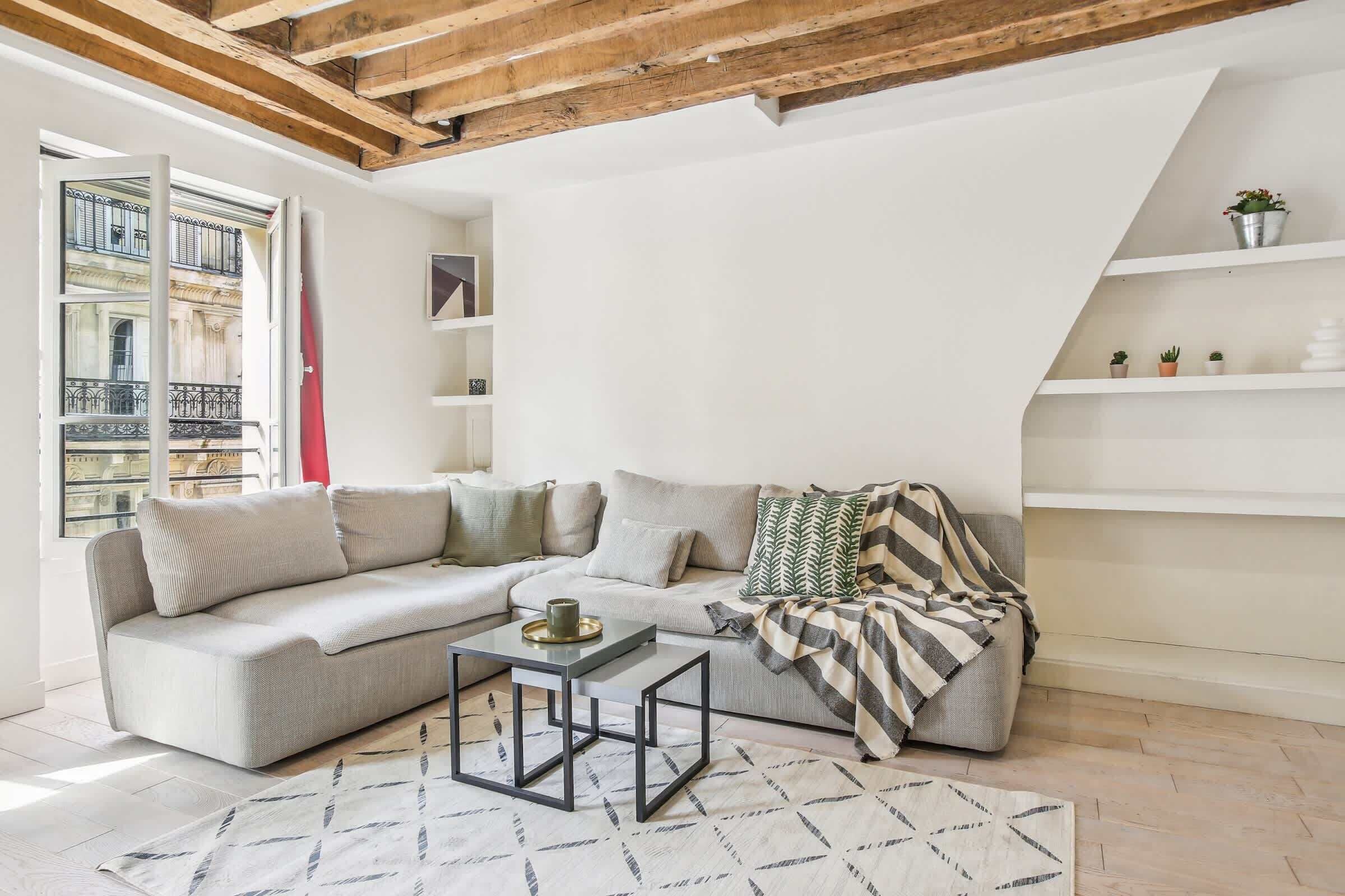 A modern living room with a grey sofa, a coffee table, and a striped throw blanket. There are white shelves with plants and a window with a view of a building.