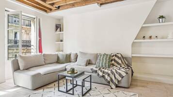 A modern living room with a grey sofa, a coffee table, and a striped throw blanket. There are white shelves with plants and a window with a view of a building.