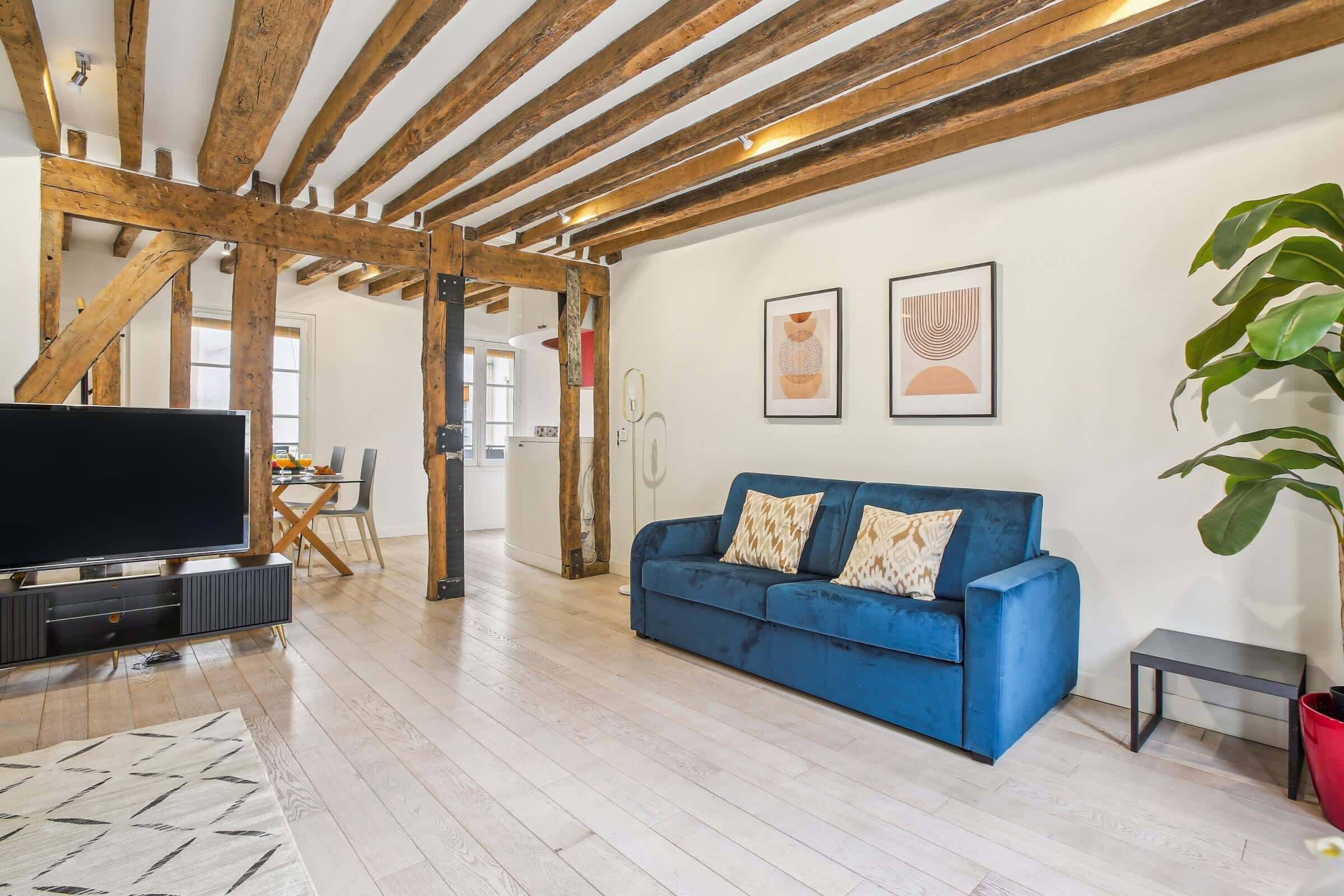 A living room with a blue sofa, wooden beams, a TV, and artwork on the wall.