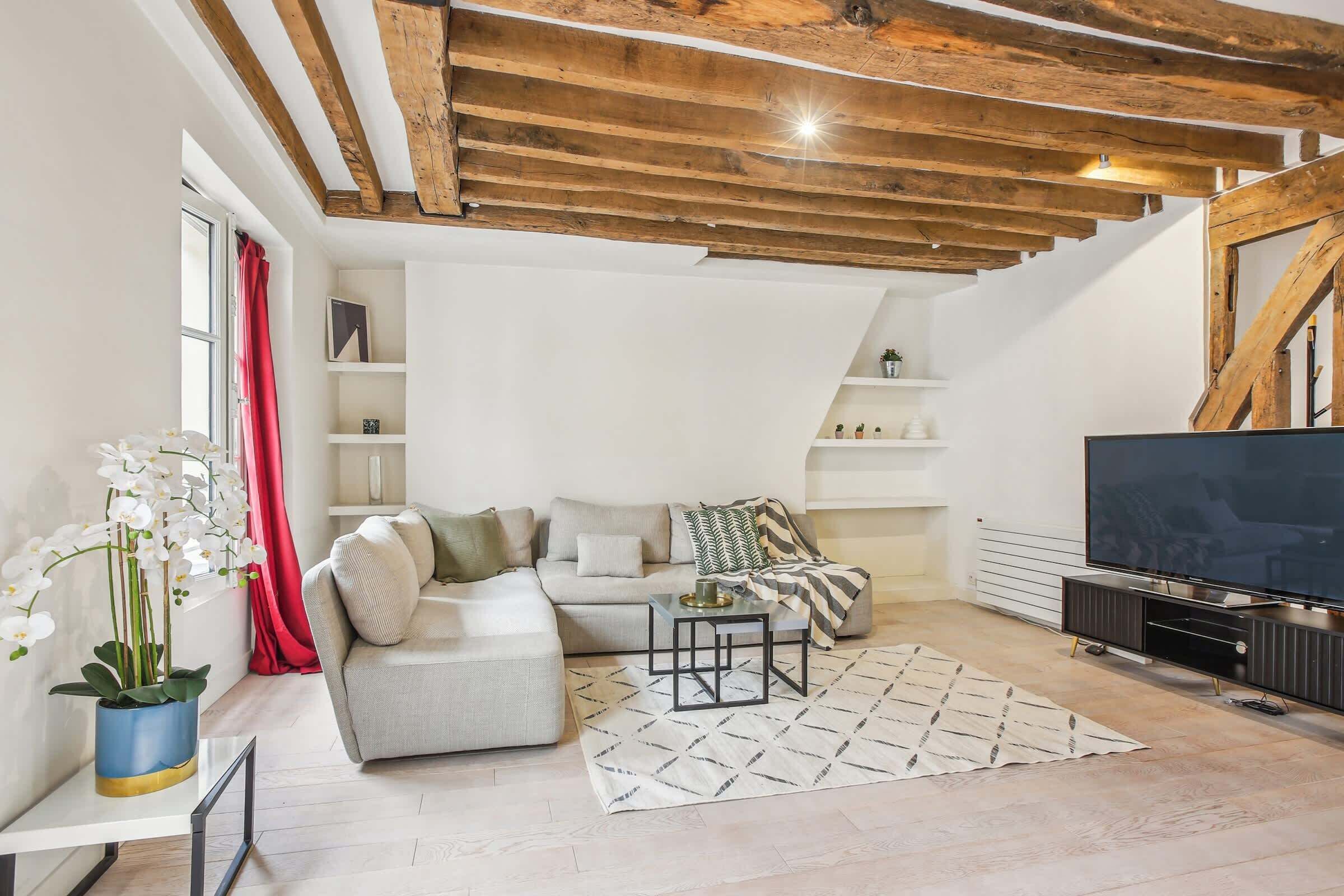 Una sala de estar moderna con un sofá modular, un televisor de pantalla plana, vigas de techo de madera y una orquídea en maceta.