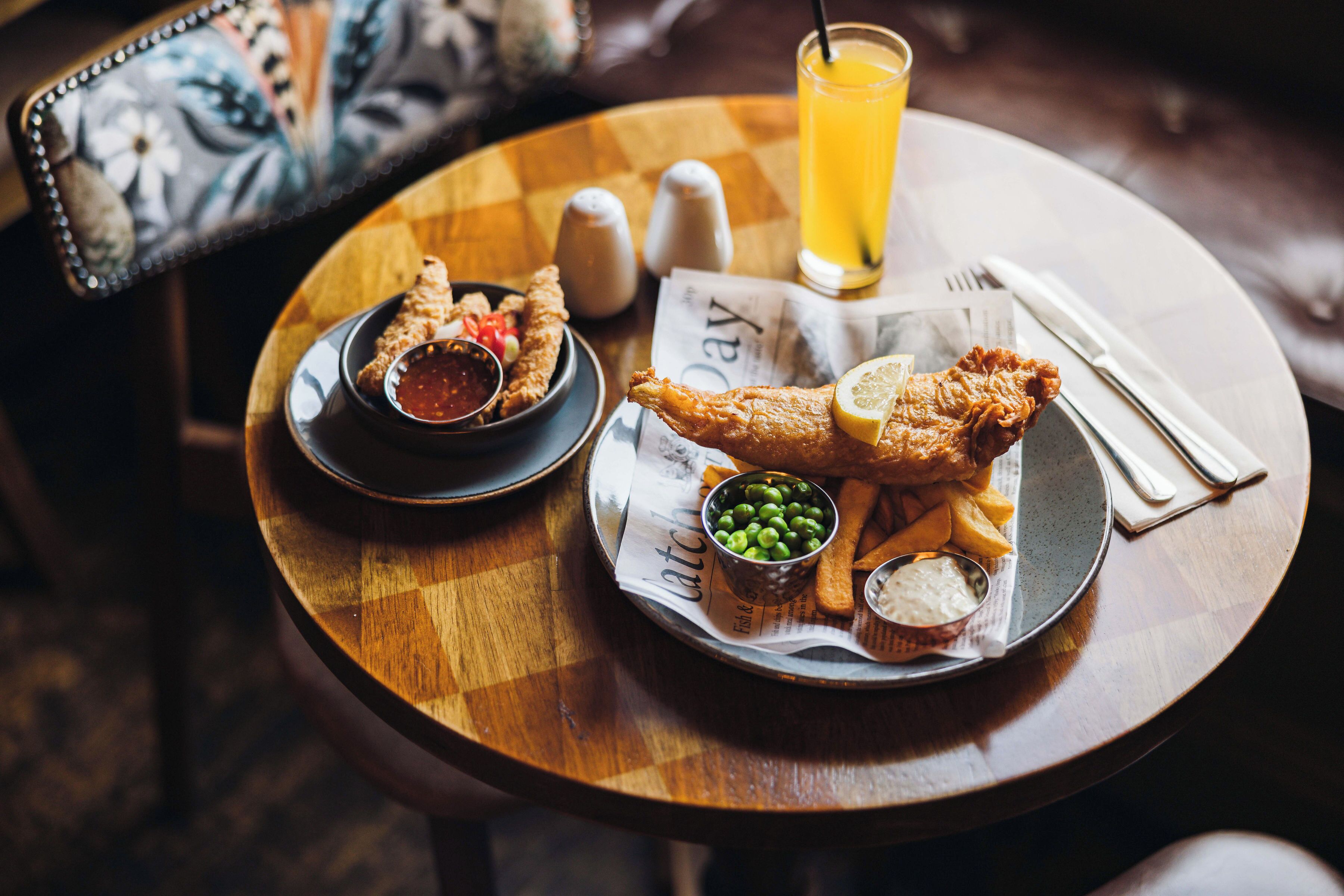 A table with a fish and chips meal, a glass of orange juice, and condiments.