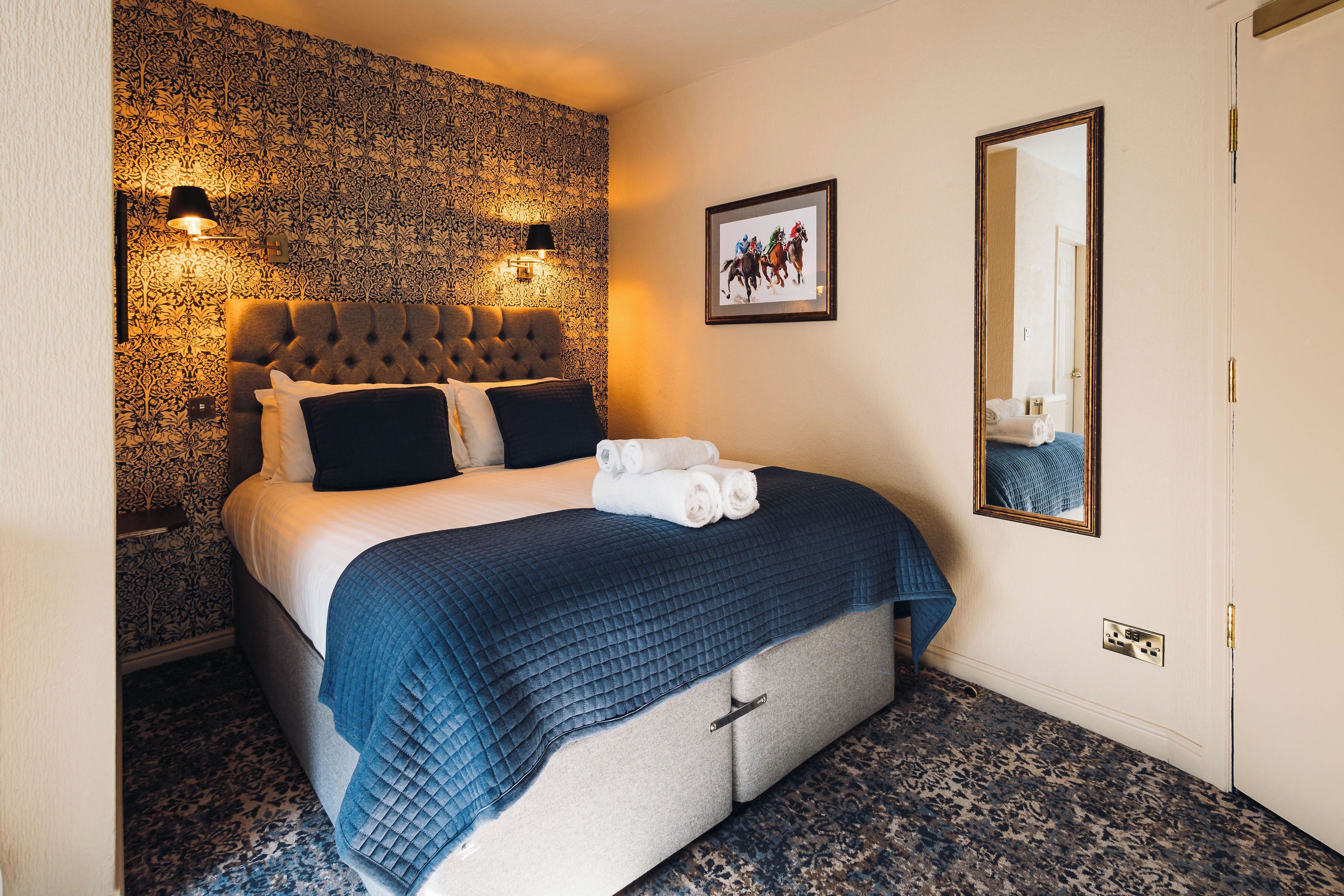 A hotel room with a large bed, a headboard with a tufted design, a blue quilted bedspread, and two wall-mounted lamps.