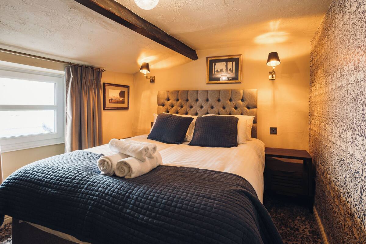 A neatly made bed with a dark quilt, two pillows, and a rolled white towel. There are two wall-mounted lamps on either side of the bed.