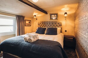 A neatly made bed with a dark quilt, two pillows, and a rolled white towel. There are two wall-mounted lamps on either side of the bed. - Richard III Hotel (Middleham)