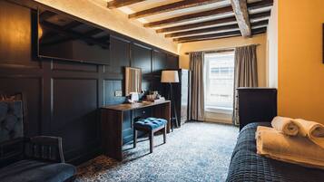 Habitación de hotel con un escritorio de madera oscura, una silla, una cama con una toalla enrollada, una lámpara y una ventana con cortinas.