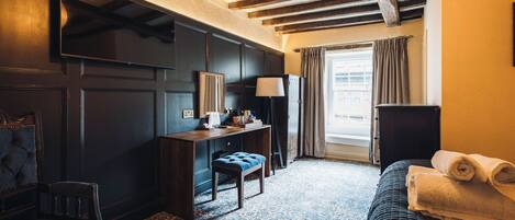 A hotel room with a dark wooden desk, a chair, a bed with a rolled-up towel, a lamp, and a window with curtains.