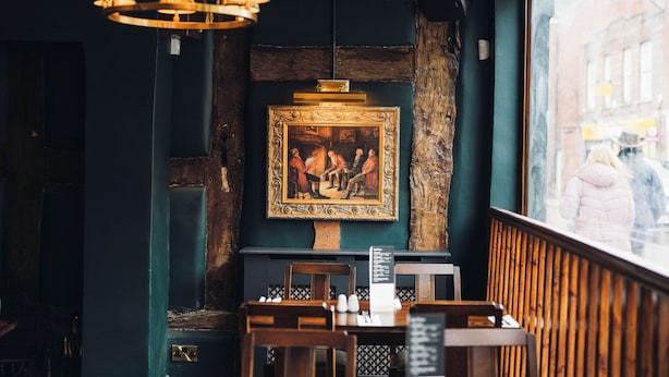 Interior de restaurante con zona de estar con bancos de madera, una araña y un cuadro histórico enmarcado en la pared.