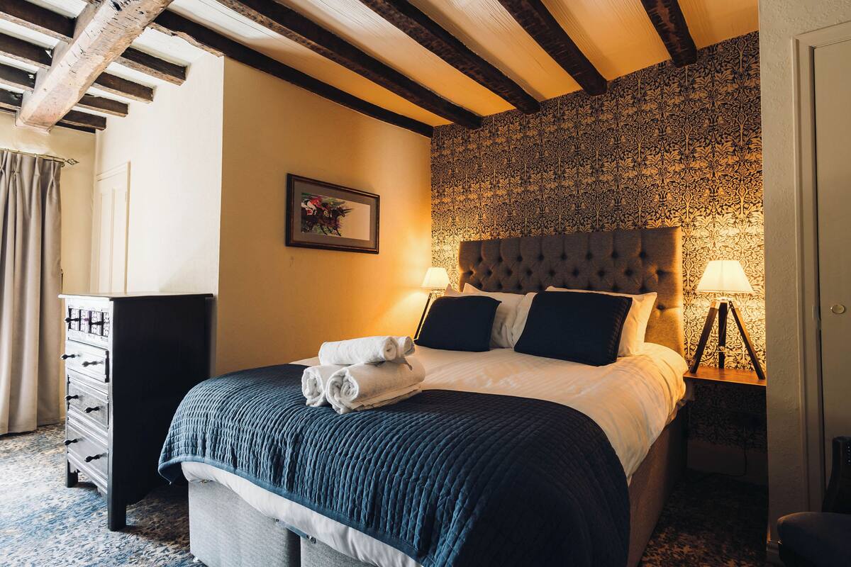 A hotel room with a bed, a dresser, a nightstand with a lamp, and a patterned wallpaper.