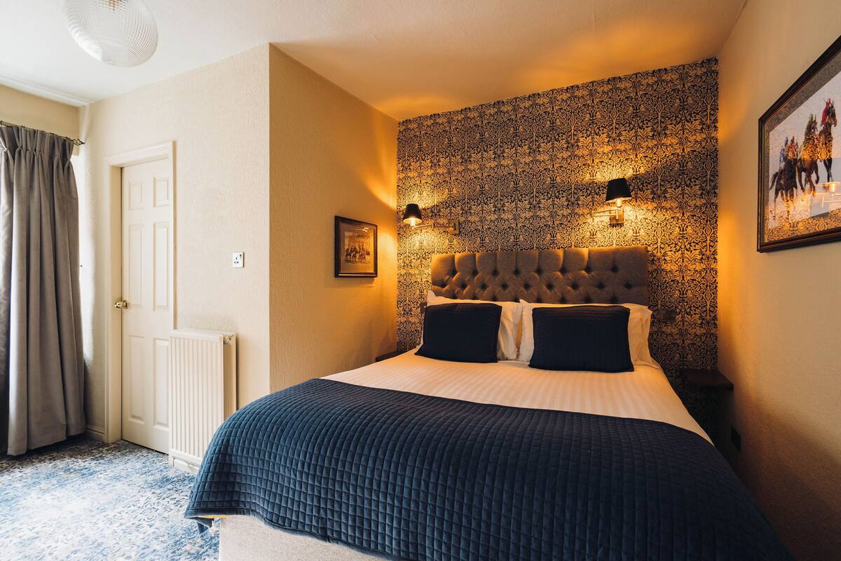 A neatly made bed with a dark quilt, a tufted headboard, and two wall-mounted reading lamps.