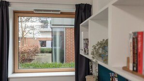 Ein Zimmer mit Fensterblick auf ein Backsteingebäude und einen Garten. Ein Bücherregal mit Büchern und eine Uhr sind an der Wand angebracht.