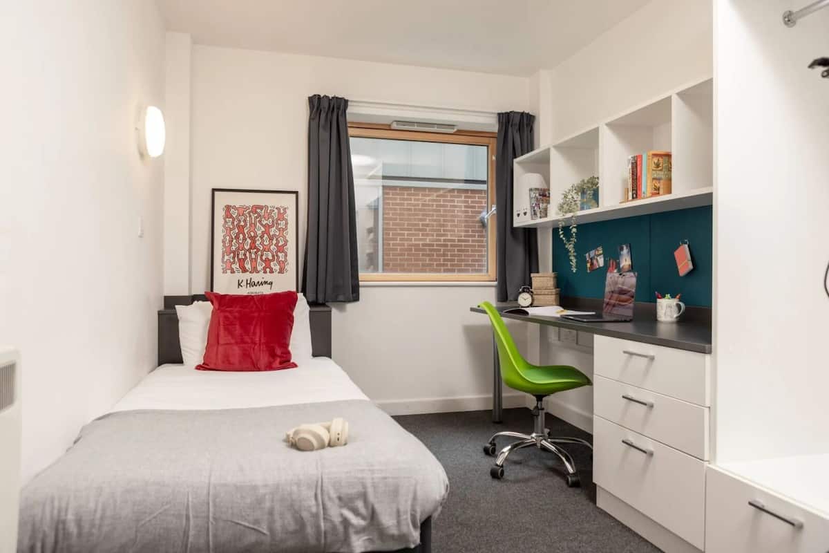 Ein gemütliches Schlafzimmer mit Bett, Schreibtisch und Stuhl. Es gibt ein Fenster mit Vorhängen, ein an der Wand befestigtes Regal mit Büchern und ein gerahmtes Bild.