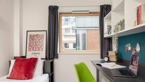 Ein Schlafzimmer mit Bett, Schreibtisch, Stuhl, Fenster, Bücherregal und einem gerahmten Bild an der Wand.