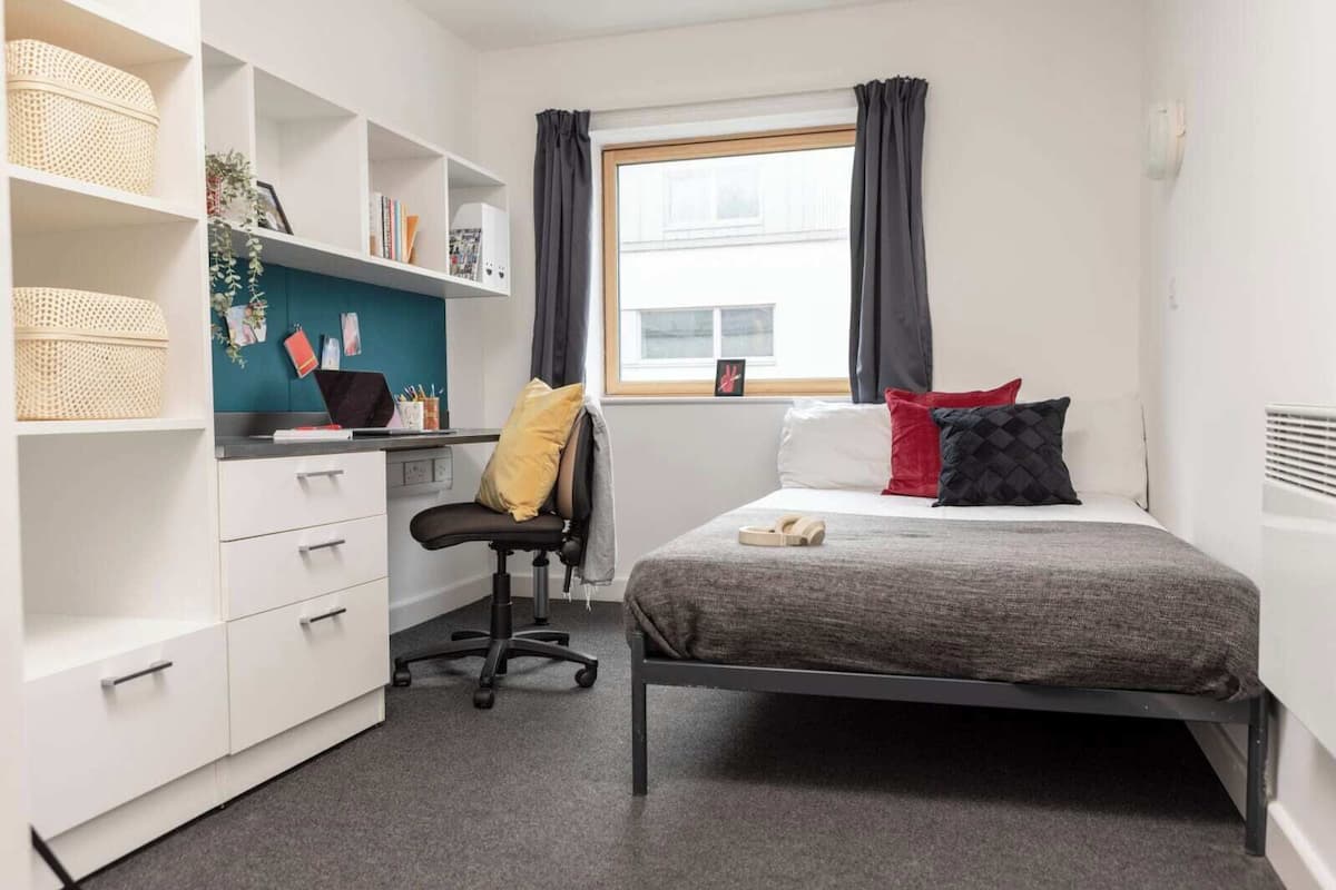 Ein gemütliches Schlafzimmer mit Bett, Schreibtisch und Stuhl. Regale mit Aufbewahrungsboxen und ein Fenster mit Vorhängen.