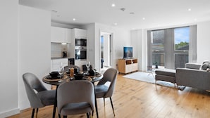 Una cocina moderna con armarios blancos, un comedor con una mesa y sillas redondas, y una sala de estar con sofá y televisor.