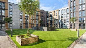 Un patio con césped artificial, una jardinera de madera con plantas y un edificio moderno con múltiples ventanas y balcones.